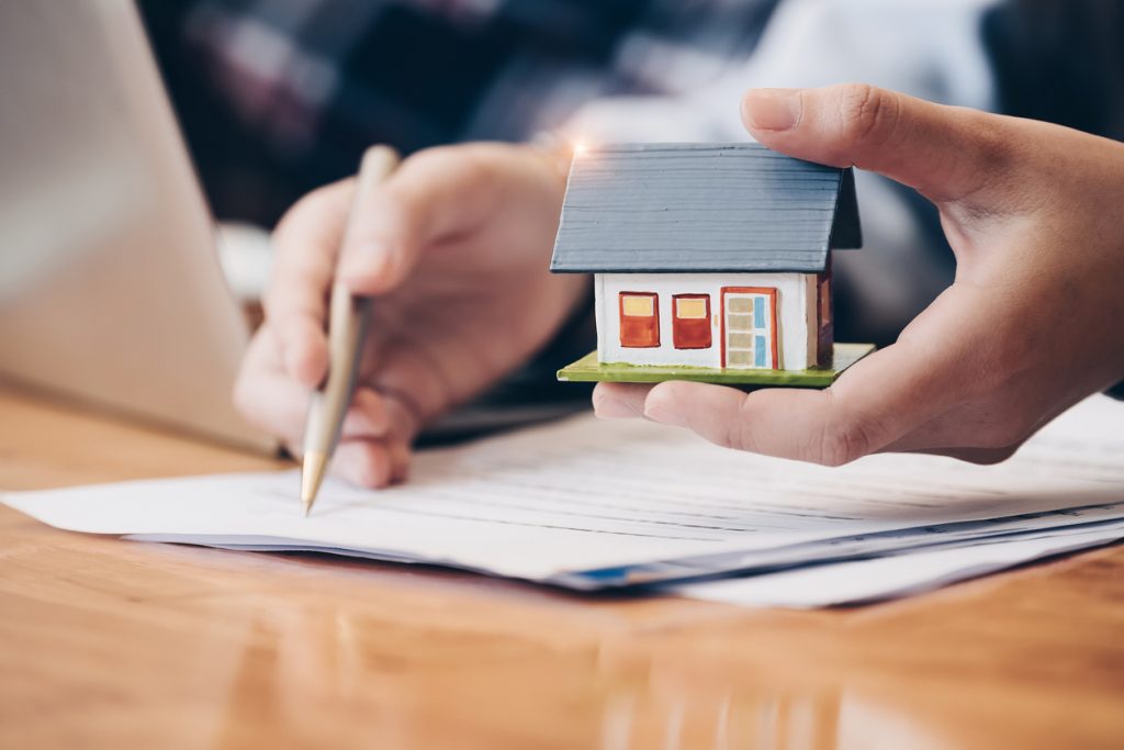 hand-holding-small-model-house-while-other-hand-holds-pencil-over-home-value-paperwork