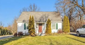 A small white house with a red door, green shutters, and evergreen trees in front.